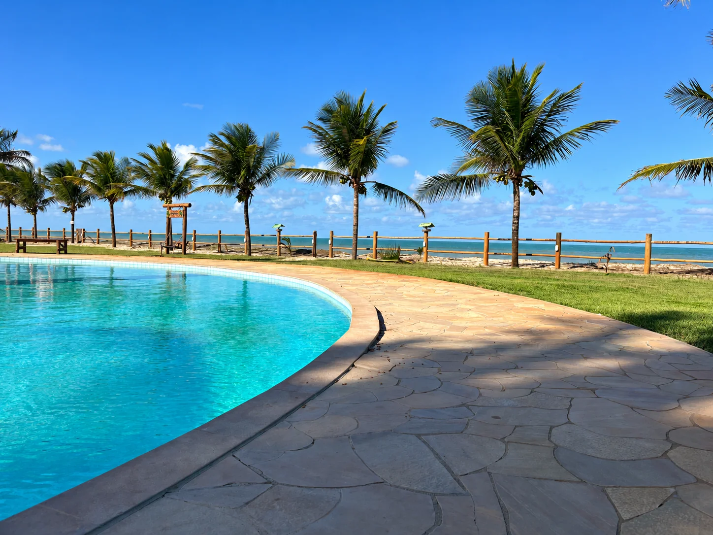 Piscina do residencial, coqueiros e mar de Alcobaça em um dia de sol